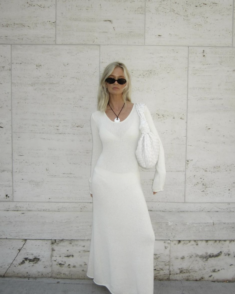 A sleek, long white dress complements the figure with its smooth fit, accessorized with a quilted white bag and dark sunglasses for a refined beach look.